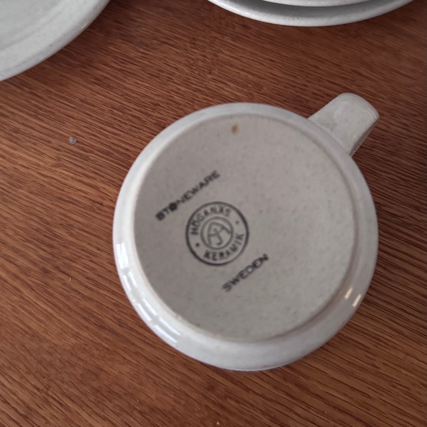 Bottom view of a Höganäs Keramik tea cup showing the stoneware mark, alongside stacked matching plates on a wooden surface.