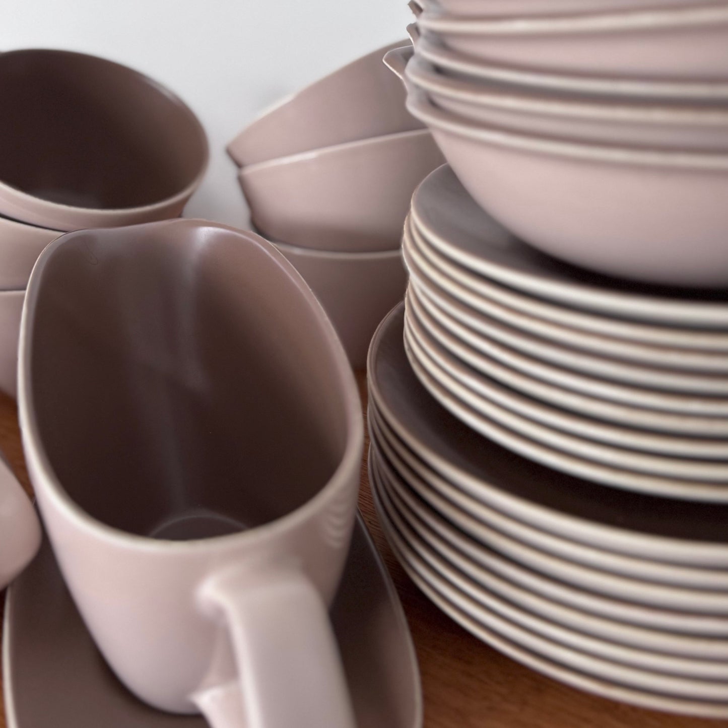 Mauve and brown Poole Pottery cups, jug, and serving dishes stacked neatly on a wooden surface
