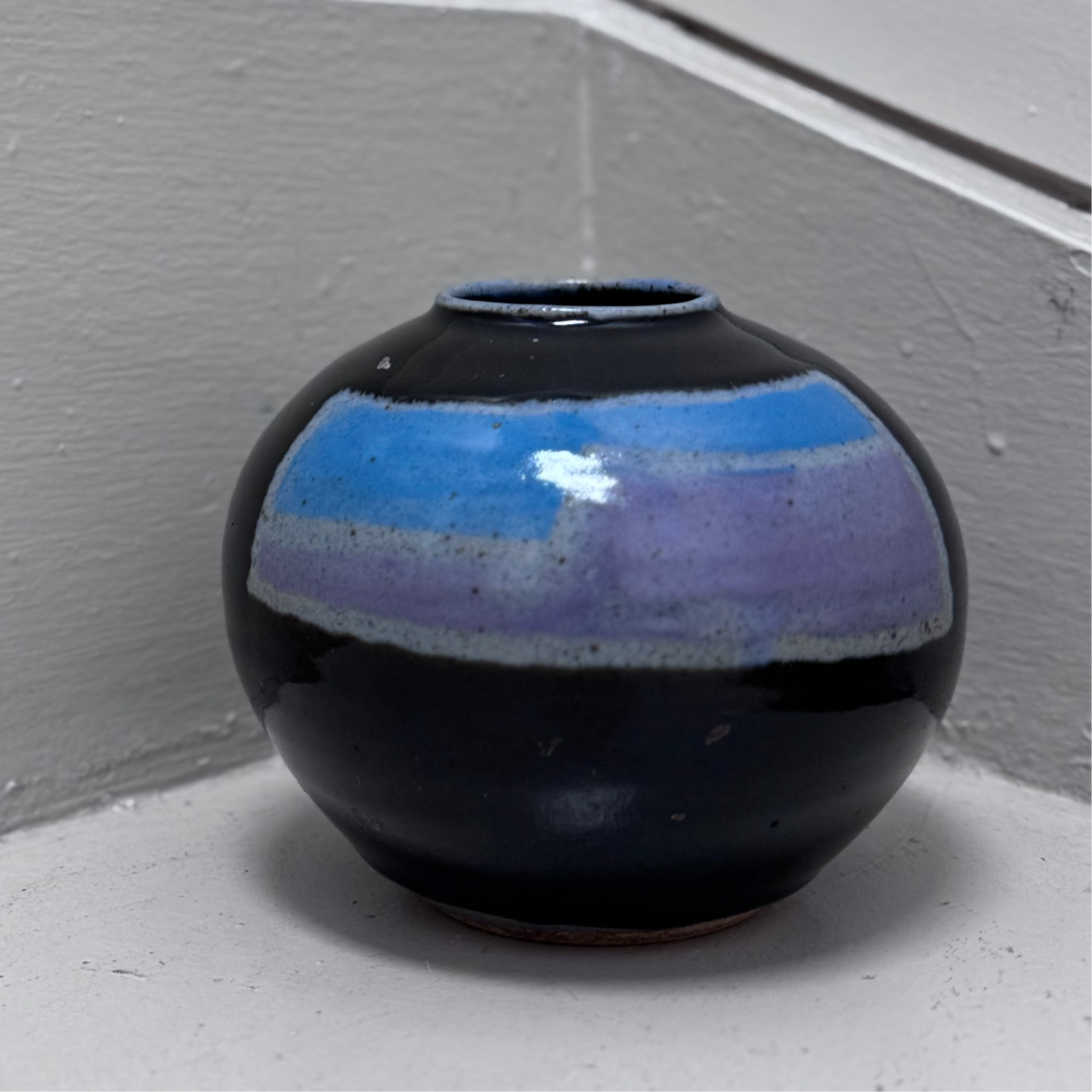 Black vase with blue top on a white surface against a gray wall.