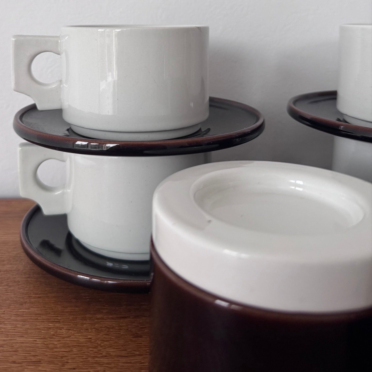 Close-up of stacked white ceramic coffee cups with modernist square handles, resting on dark brown saucers with a glossy finish.
