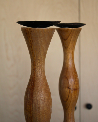 Two handcrafted wooden candlestick holders with a flared top and dark ceramic candle cups, set against a light wood paneled background.