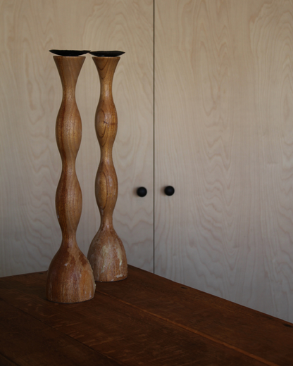 Two tall, twisted wooden candlesticks with a natural light brown finish, standing on a dark wooden table in front of light wood paneled doors.