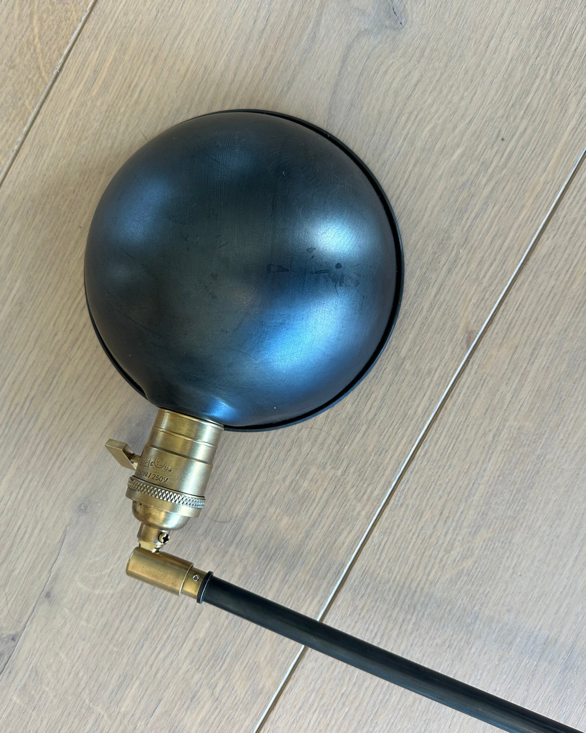 A vintage-style industrial floor lamp with a large black spherical shade, brass socket, and black cord, photographed from above on a light wood plank floor.