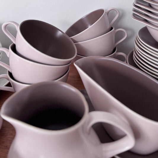 Mauve and brown Poole Pottery cups, jug, and serving dishes stacked neatly on a wooden surface