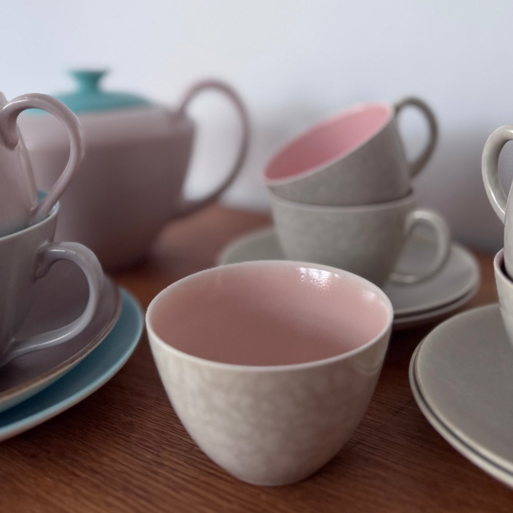Assorted ceramic cups and saucers in grey and pastel pink with a teal-lidded teapot on a wooden surface