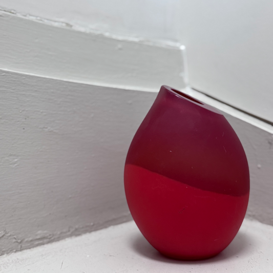 Modern red frosted glass vase with a rounded shape and tilted rim, placed on a white corner step