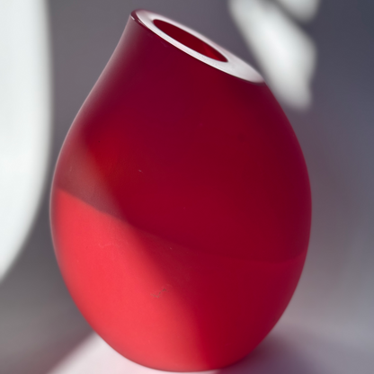 Bold red frosted glass vase with a curved shape and asymmetrical rim, set against a softly lit background