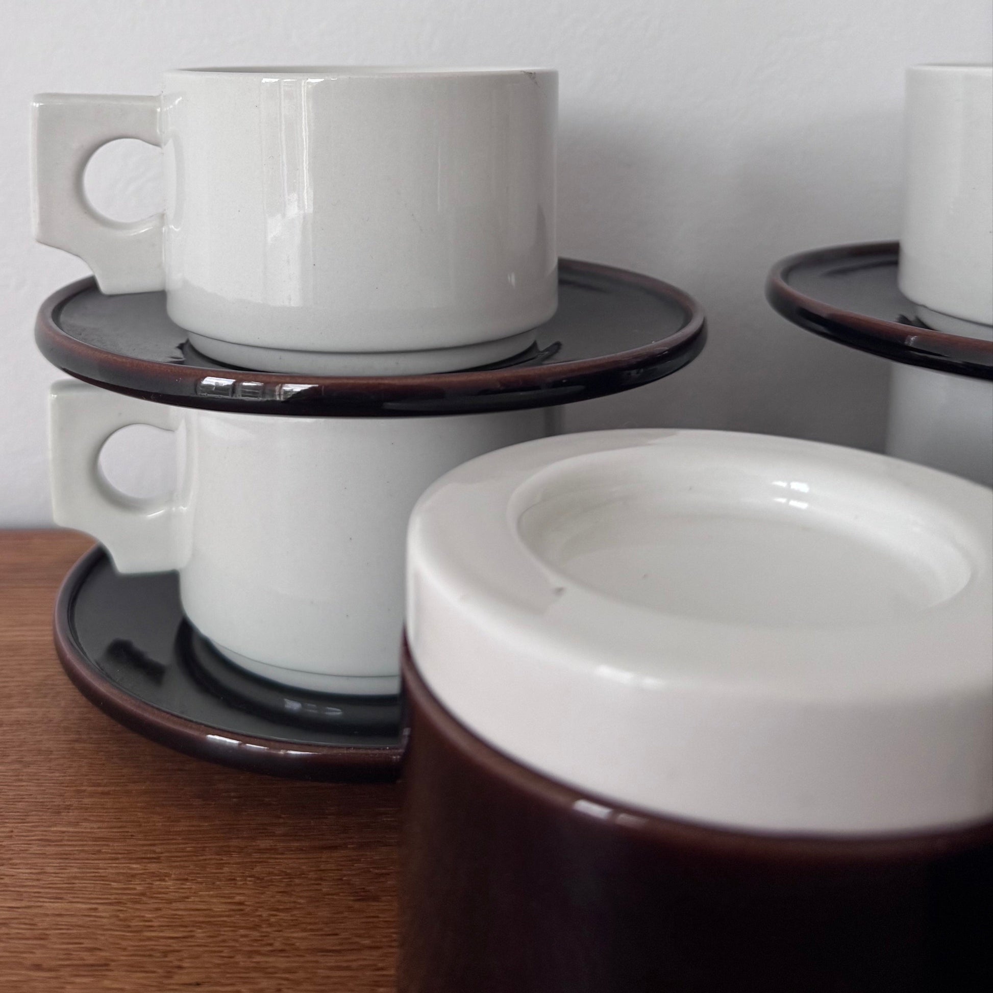 Close-up of stacked white ceramic coffee cups with modernist square handles, resting on dark brown saucers with a glossy finish.