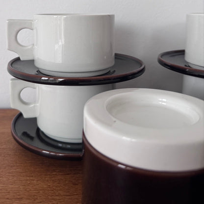 Close-up of stacked white ceramic coffee cups with modernist square handles, resting on dark brown saucers with a glossy finish.