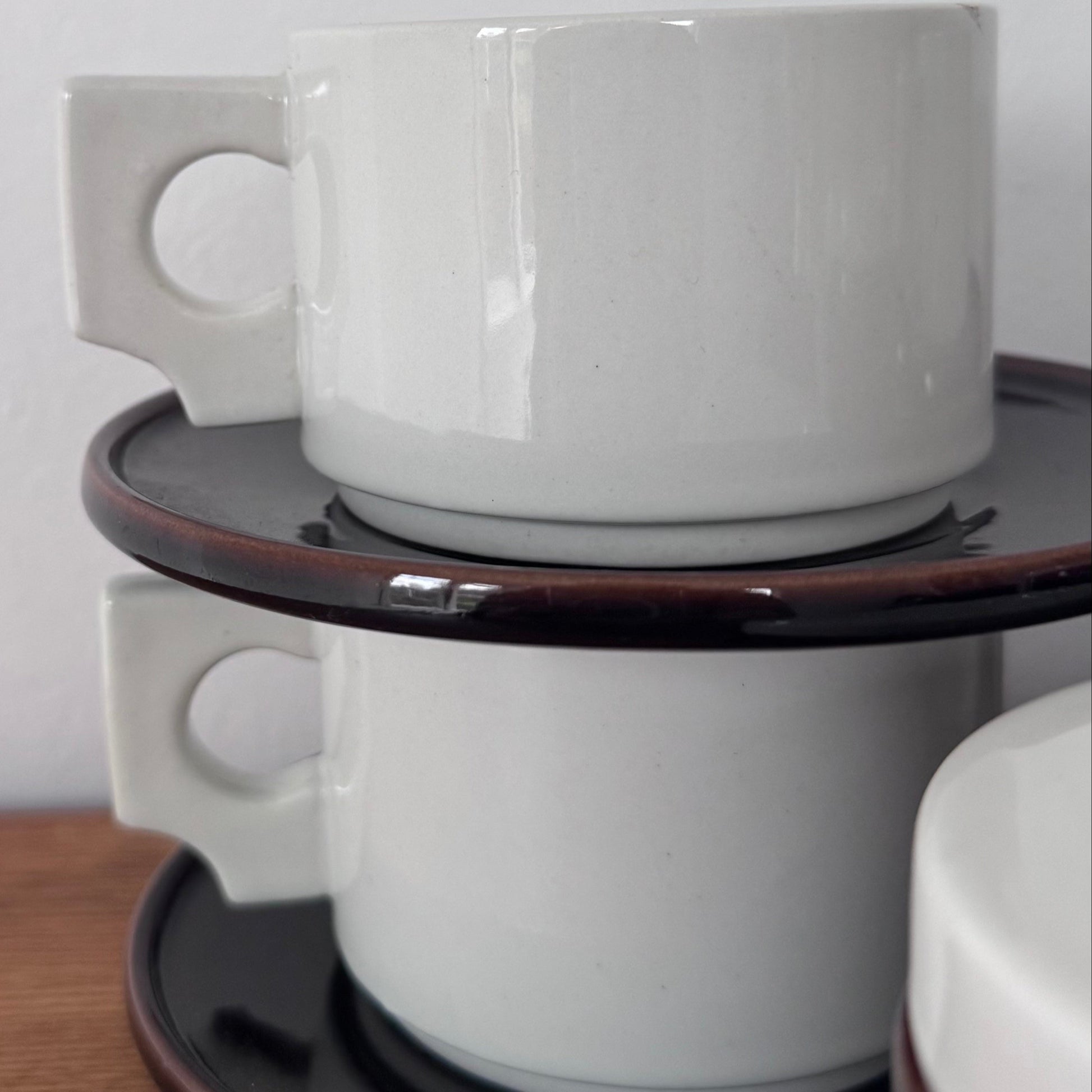 Close-up of stacked white ceramic coffee cups with modernist square handles, resting on dark brown saucers with a glossy finish.