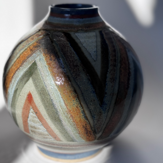 A round New Zealand studio pottery vase featuring bold zig-zag patterns in earthy tones of green, orange, black, blue, and cream, with a glossy finish.