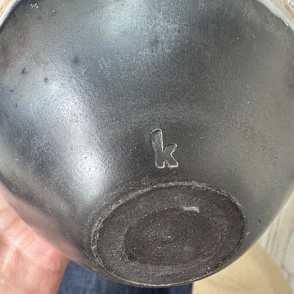 Underside of a ceramic pot with a smooth black base, featuring a carved 'K' maker’s mark near the center.